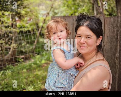 Mamma che tiene figlia e guarda la fotocamera in cortile naturale Foto Stock