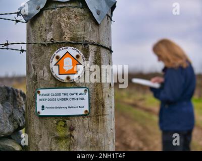 La segnaletica segna la strada per il Pennine Bridleway National Trail su un palo di legno intemperiato. Una sola donna consulta una mappa sullo sfondo. Foto Stock