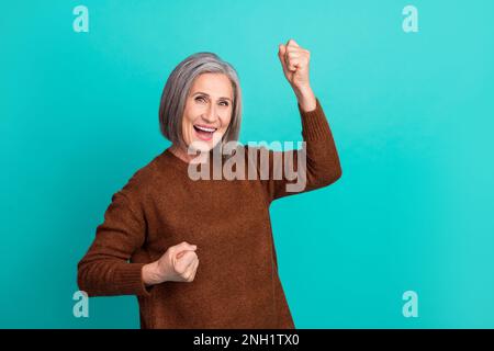 Foto di donna allegra vestito marrone trendy alzare le mani pugno grida hooray sì isolato su sfondo di colore ciano Foto Stock