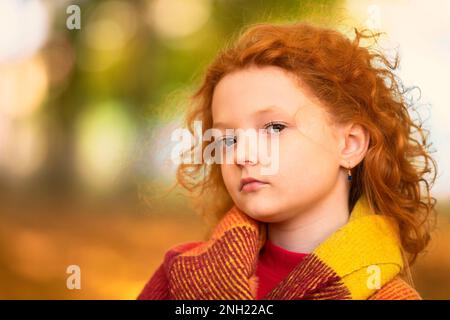 Una ragazza dai capelli rossi si avvolse in una coperta di lana in un parco autunnale. Bambino in autunno. Foto Stock