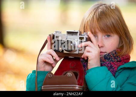 Piccola ragazza dai capelli rossi con una macchina fotografica retrò nel parco autunnale. Fotografo bambino. Foto Stock