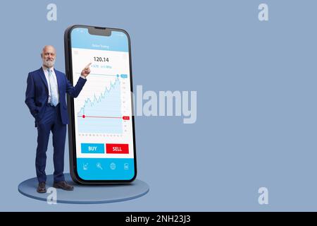 Uomo d'affari accanto a un grande smartphone con app di trading online, sta sorridendo e puntando sullo schermo, concetto di trading online Foto Stock