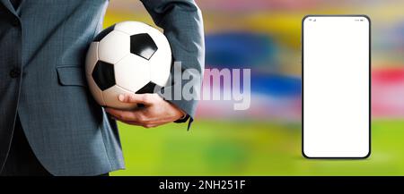 Manager che tiene una palla di calcio e app per smartphone, campo di calcio in background, concetto di app sportive e mobili Foto Stock