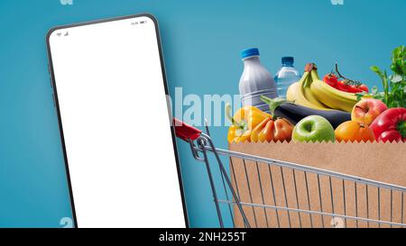 Supermercato carrello pieno di generi alimentari e smartphone con schermo vuoto, concetto di shopping online drogheria Foto Stock