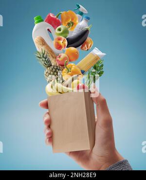 Cliente che tiene una borsa di carta in miniatura con alimentari freschi che cadono all'interno, concetto di shopping della drogheria Foto Stock