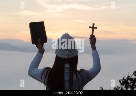 Le mani della giovane donna che tengono la Sacra Bibbia e sollevano la croce cristiana con il tramonto chiaro sfondo. Foto Stock