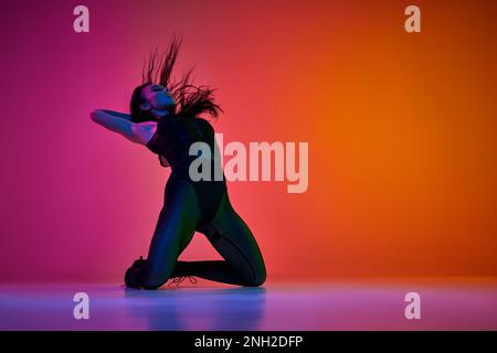 Auto-espressione attraverso i movimenti. Giovane donna che danza alto tallone dance stile su gradiente rosa arancio sfondo studio in luce al neon Foto Stock