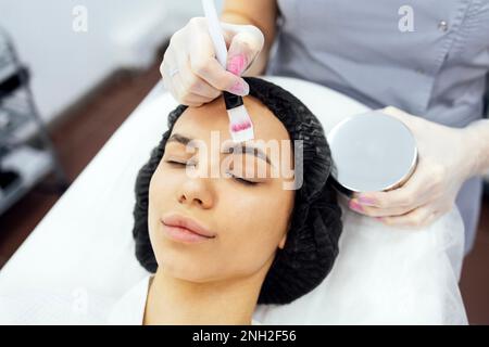 Top view ritratto di una bella ragazza tranquilla in un accappatoio bianco sta facendo una procedura cosmetica. Il maestro di bellezza applica una maschera ringiovanente al cl Foto Stock