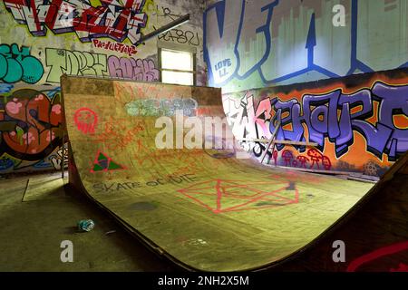 Una grande rampa di skateboard in compensato illuminata dal sole circondata da pareti ricoperte di luminosi graffiti in un vecchio edificio idro abbandonato. Foto Stock