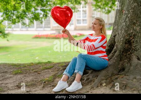 una donna in jeans che tiene un palloncino rosso sotto l'albero nel parco Foto Stock