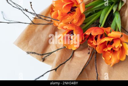 Un bellissimo bouquet primaverile di tulipani di arance peony, insieme a rami di ciliegie giovani, si trova in carta artigianale su un tavolo bianco Foto Stock