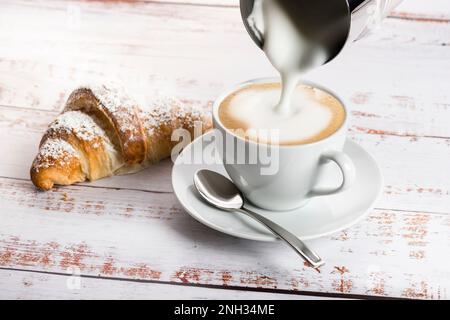 Preparare il cappuccino con il pannarello su un tavolo di legno con croissant. Foto Stock
