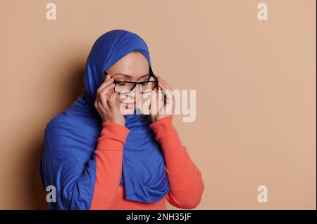 Elegante donna musulmana con testa coperta in hijab, mette su occhiali eleganti su sfondo crema isolato con spazio copia Foto Stock