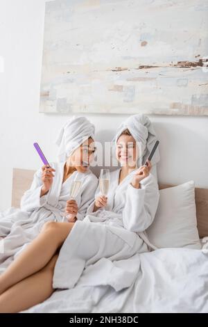 donna afro-americana stupita con allegro amico in possesso di champagne e lime unghie mentre si siede in bianco accappatoi e asciugamani a letto, immagine stock Foto Stock