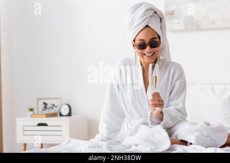 donna afro-americana felice in accappatoio spugna e occhiali da sole alla moda seduta sul letto con un bicchiere di champagne, immagine stock Foto Stock