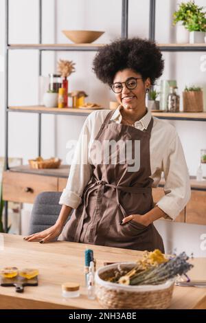 donna afro-americana felice con mano in tasca di grembiule in piedi vicino al tavolo con saponi sfocati ed erbe essiccate, immagine di scorta Foto Stock