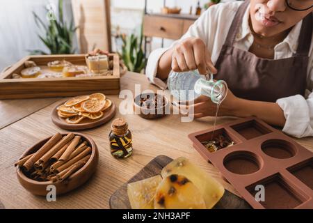 vista parziale dell'artigiano afro-americano che versa sapone liquido in silicone stampo vicino spezie e cannella bastoncini con fette d'arancia essiccata, immagine stock Foto Stock