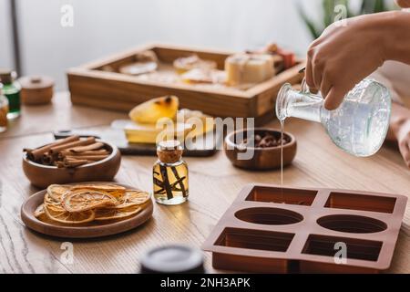 vista parziale di afro-americano artigianale versando sapone in silicone stampo vicino a olio essenziale e fette d'arancia essiccata, immagine stock Foto Stock