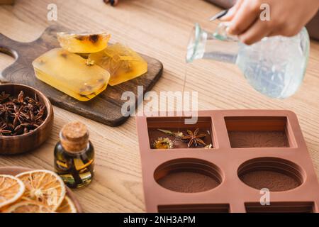 vista parziale dell'artigiano afro-americano che versa sapone liquido in stampo di silicone con erbe essiccate vicino all'olio essenziale e fette di arancia, immagine stock Foto Stock