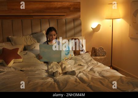 Piccola ragazza lettura libro in camera da letto illuminato da lampada da notte Foto Stock