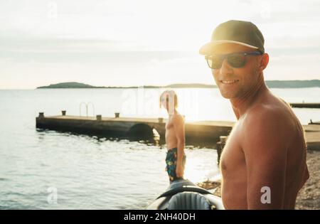 Sorridente uomo di mezza età in occhiali da sole e berretto da baseball eleganti guardando la fotocamera. Padre con figlio adolescente che gode di una bella luce al tramonto sul mare. Fa Foto Stock