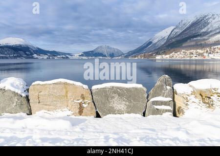 Un tranquillo ambiente invernale mette in mostra rocce innevate accanto a un lago fermo, incorniciate da maestose montagne sotto un cielo nuvoloso. Foto Stock