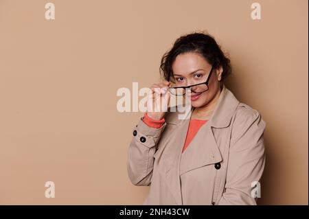 Donna sorridente multi-etnica positiva di mezza età in abbigliamento casual, indossa occhiali eleganti, guardando la fotocamera su sfondo beige isolato con libero Foto Stock