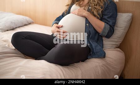 Donna incinta seduta sul letto che tiene la pancia Foto Stock