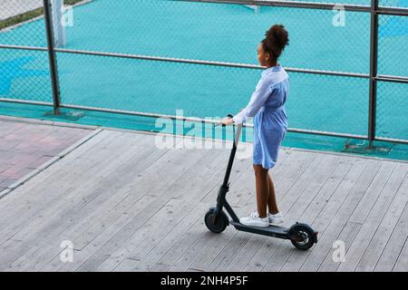 Ragazza afro-americana adolescente in abito casual e scarpe sportive in sella scooter elettrico, mentre si muove lungo il filo di ferro rete recinto Foto Stock
