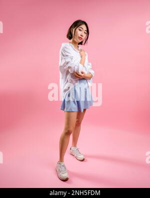 Full Body Asian Woman Short Blue gonna White Blouse sfondo rosa in piedi con braccia avvolte intorno vita e torace Vista laterale Foto Stock