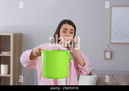 Giovane donna che chiama il servizio di riparazione del tetto mentre raccoglie l'acqua che fuoriesce dal soffitto a casa Foto Stock