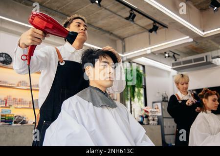 Giovane parrucchiere asiatico maschio che indossa grembiule asciugando bei capelli di giovane uomo in un moderno barbiere mentre si concentra sul lavoro Foto Stock