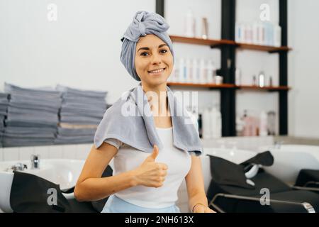 Ritratto di bella giovane donna caucasica in salone con i pollici gesto di segno in su mentre avendo avvolto un asciugamano sopra bagnato capelli dopo il lavaggio dei capelli Foto Stock