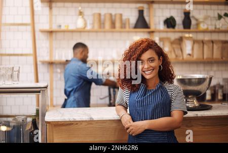 Oggi il buon umore è sponsorizzato dal caffè. una giovane e attraente donna d'affari in piedi nel suo caffè, mentre un barista lavora dietro di lei. Foto Stock