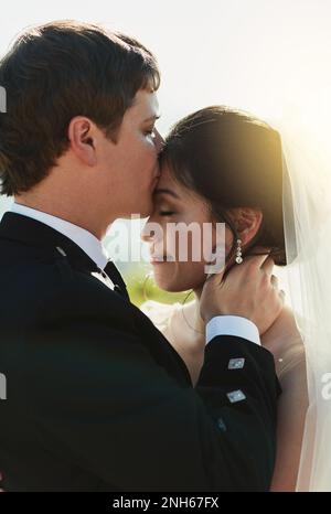 Finalmente un po' di tempo da solo. un giovane sposo allegro che dà alla sua sposa un bacio sulla fronte mentre si stanno tenendo l'un l'altro fuori. Foto Stock