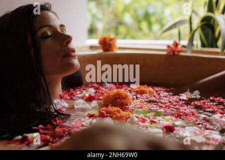 Foto di una bella giovane donna che si rilassa nella vasca da bagno con petali di fiori. Donna con bagno di fiori in una spa. Foto Stock