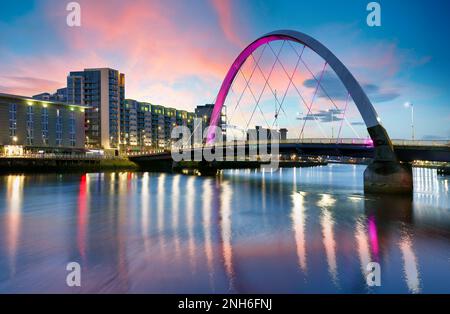 Bellissimo Sunset Clyde Arc Bridge attraverso il fiume a Glasgow, Scozia, Regno Unito. E' bel tempo con riflessi sull'acqua, il cielo blu, le luci degli edifici Foto Stock