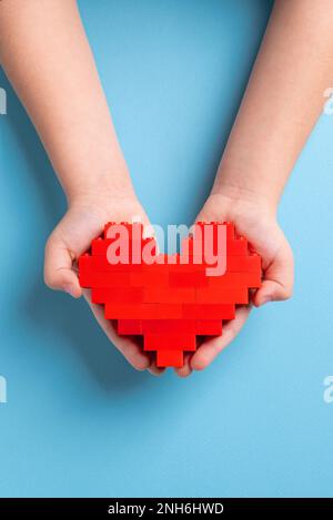 Mani di bambina che tiene il cuore fatto di mattoni di plastica su sfondo blu Foto Stock