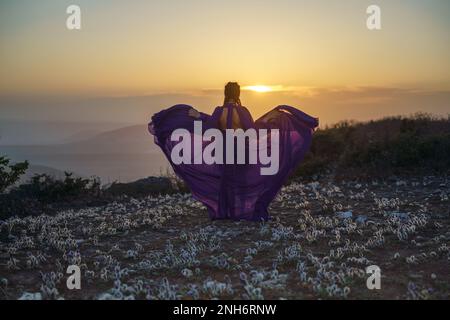 Tramonto viola abito donna montagne. Ascesa del mistico. tramonto sulle nuvole con una ragazza in un lungo vestito viola. Nel prato c'è un prato Foto Stock