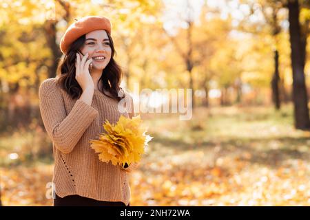 Bella giovane donna felice nel parco autunnale parlando su uno smartphone all'aperto in alberi gialli luminosi in autunno. Colorato concetto autunnale. Spazio di copia Foto Stock