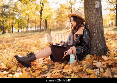 Giovane donna d'affari che lavora sul suo notebook nel parco. Donna freelance seduta sul fogliame sotto l'albero utilizzando un computer portatile per il lavoro. Spazio di copia Foto Stock