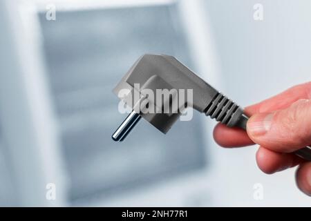 Spina elettrica in mano sul fondo del riscaldatore alogeno della casa fredda in sfocatura. Apparecchio di riscaldamento scollegato. Simbolo del risparmio di energia elettrica dovuto all'e Foto Stock