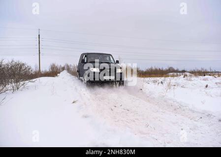 Il SUV russo 'UAZ Patriot' procede rapidamente su una strada innevata nel campo in inverno sullo sfondo della foresta aggrappata alla ruota della tempesta di neve. Foto Stock