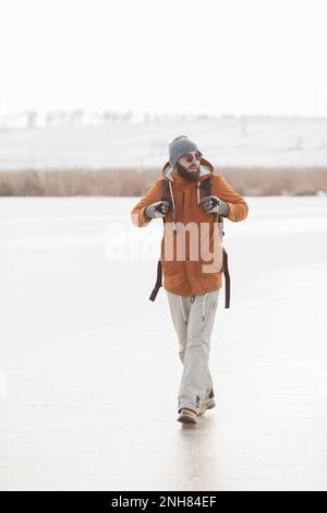 Un uomo bearded con uno zaino in abiti caldi di inverno sta camminando su un lago ghiacciato in inverno. Il concetto di trekking e di stile di vita attivo. Foto Stock