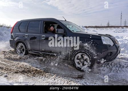 Il crossover fuoristrada "Nissan X-Trail 4x4" solleva rapidamente la neve in inverno con una donna allegra passeggero che guarda fuori dalla finestra aperta. Foto Stock