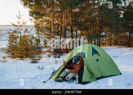 Giovane uomo beared sorridente, bevendo caffè e prendendo selfie in montagna d'inverno vicino tenda. Viaggiatore maschio con barba in un cappello e una giacca calda prendere Foto Stock