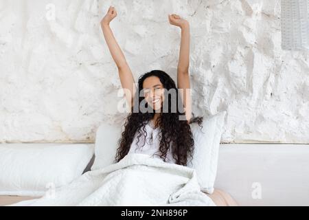 Bruna donna caucasica felice millenaria con lunghi capelli svegliarsi, seduta su morbido letto, stretching corpo Foto Stock