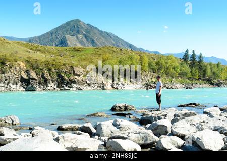 Maschio pescatore in pantaloni roll up, t-shirt bianca e berretto pesca filatura caldo luminoso giorno veloce turchese montagna fiume Katun nella Altai sulla pietra Foto Stock