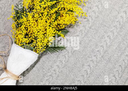 Bouquet con rami di mimosa su coperta grigia lavorata a maglia. Tessera vacanze con fiori di mimosa. Composizione primaverile adatta per il giorno della madre o per il giorno della donna. COP Foto Stock