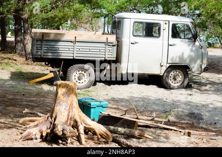 Russian pick - up- camion 'UAZ 3303 tadpole' è accanto con un'ascia bloccata in un ceppo sulla spiaggia di sabbia nella foresta tra gli alberi in Altai Foto Stock